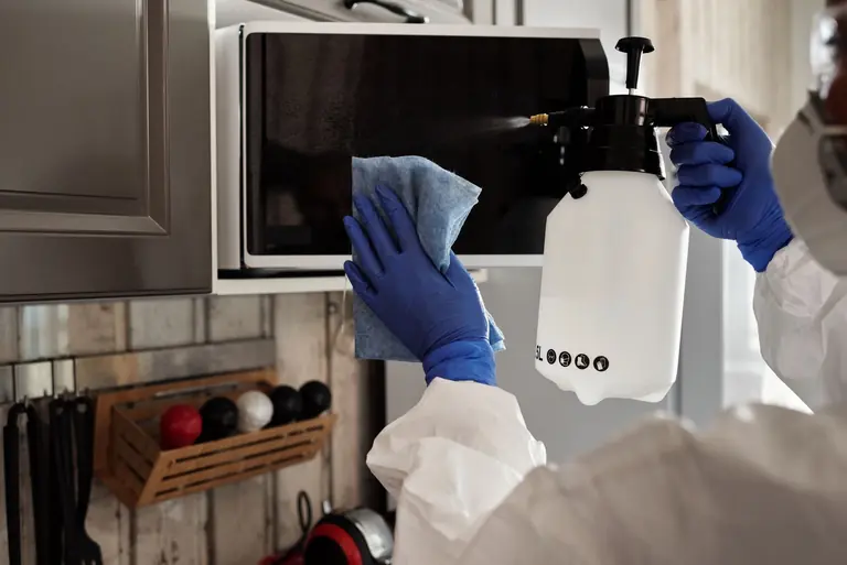 Individual in protective gear cleaning microwave using spray bottle and cloth indoors.